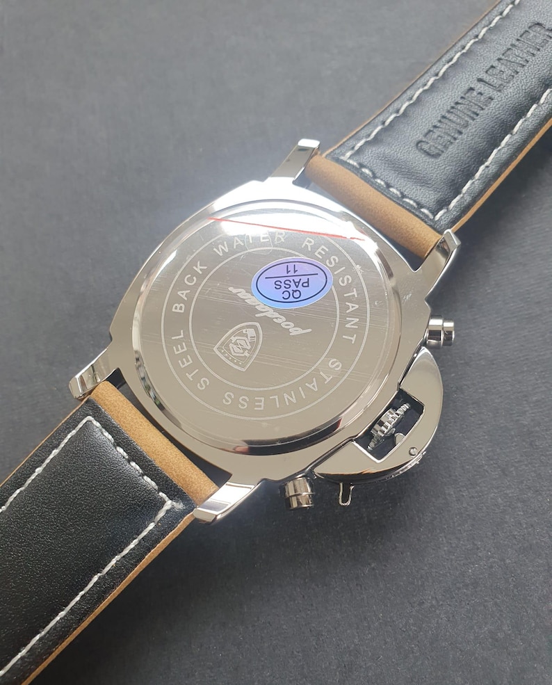 May include: The back of a silver watch with a black leather band. The watch face says "Water Resistant" and "Stainless Steel Back". There is a blue sticker on the watch that says "QC Pass 11".