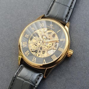 May include: A gold-tone skeleton watch with black Roman numeral hour markers and a black leather band. The watch face is transparent, revealing the intricate inner workings of the watch.