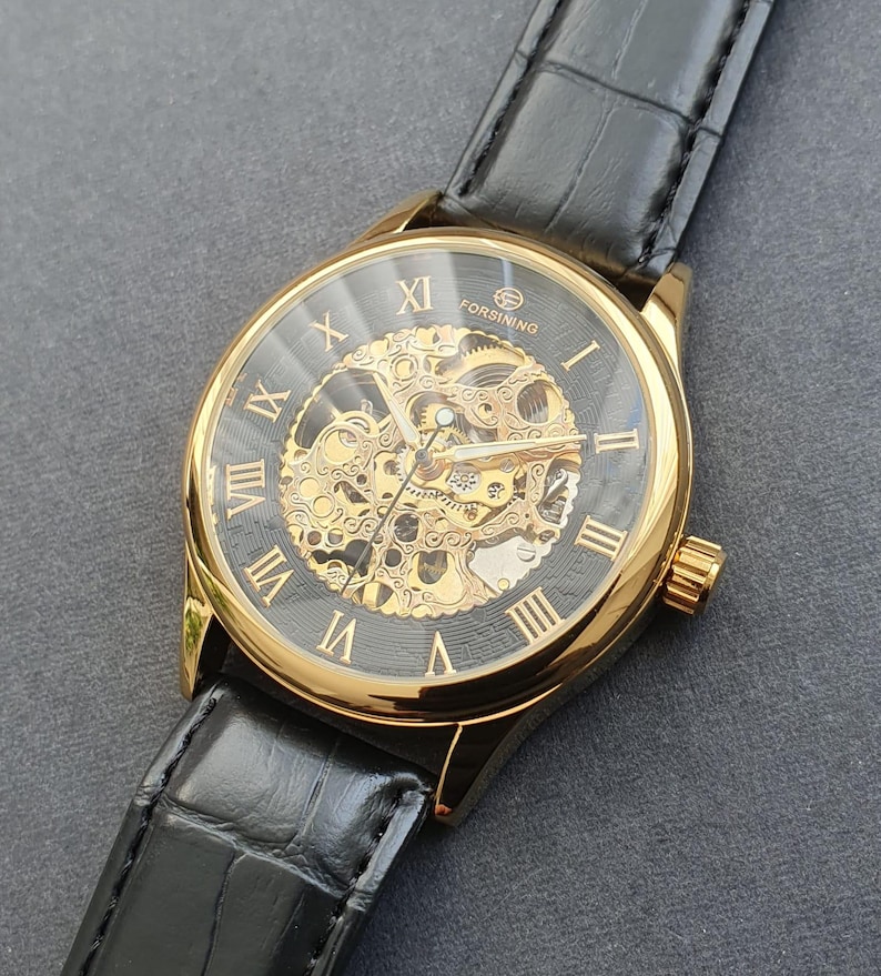 May include: A gold-tone skeleton watch with black Roman numeral hour markers and a black leather band. The watch face is transparent, revealing the intricate inner workings of the watch.