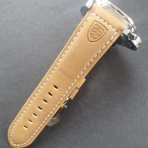 May include: A tan leather watch band with white stitching and a silver buckle. The band has a logo embossed on it.