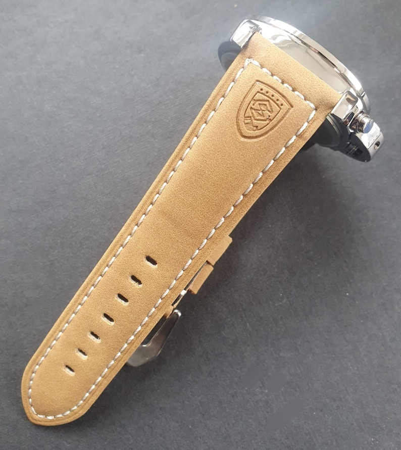 May include: A tan leather watch band with white stitching and a silver buckle. The band has a logo embossed on it.
