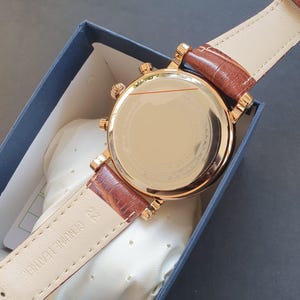 May include: A gold-tone watch with a brown leather band. The watch has a round face and a chronograph function. The back of the watch is visible, showing the watch's mechanism.