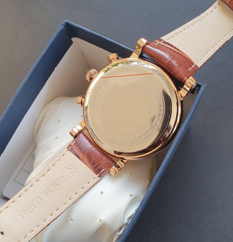 May include: A gold-tone watch with a brown leather band. The watch has a round face and a chronograph function. The back of the watch is visible, showing the watch's mechanism.