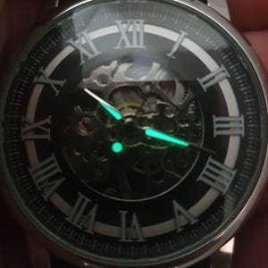 Puede incluir: Un reloj negro y plateado con n&uacute;meros romanos en la esfera. El reloj tiene un dise&ntilde;o de esqueleto, que permite ver el mecanismo interno. Las manecillas del reloj brillan en verde en la oscuridad.