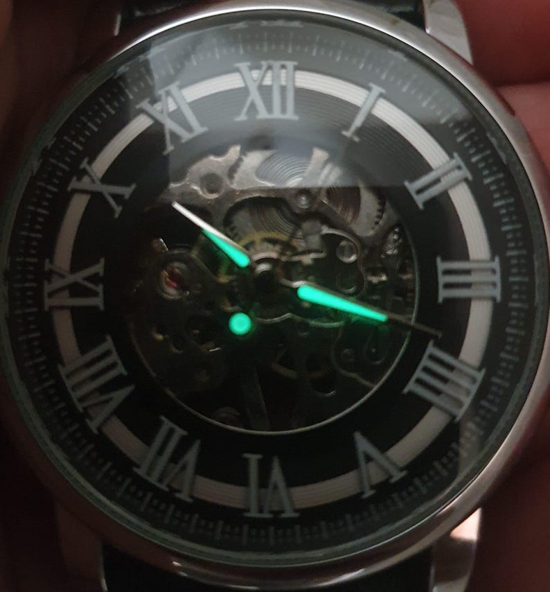Puede incluir: Un reloj negro y plateado con n&uacute;meros romanos en la esfera. El reloj tiene un dise&ntilde;o de esqueleto, que permite ver el mecanismo interno. Las manecillas del reloj brillan en verde en la oscuridad.