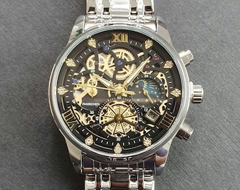 Peut inclure: Une montre automatique argentée et dorée avec un cadran noir et un design squelette. La montre possède un guichet de date, un indicateur de phase lunaire et un sous-cadran 24 heures. La montre est ornée de petits diamants autour de la lunette.