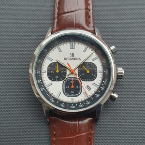 Op de afbeelding: Een zilveren en witte chronograaf polshorloge met een bruin leren bandje. De wijzerplaat van het horloge heeft zwarte markeringen en een rode secondewijzer. De merknaam "INCASEDA" is op de wijzerplaat van het horloge gedrukt.