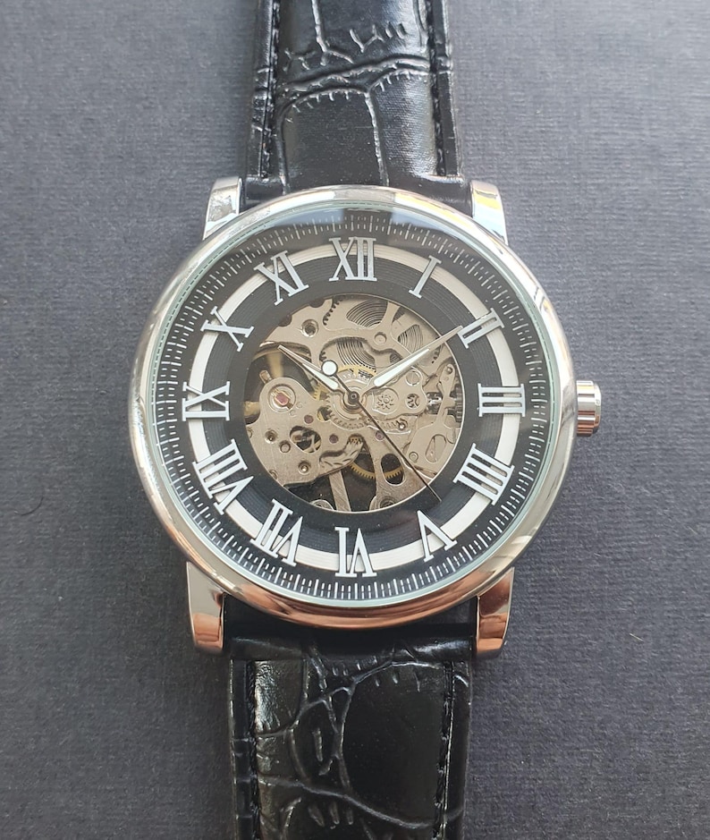 Puede incluir: Un reloj de pulsera plateado con una esfera negra y marcadores de hora en n&uacute;meros romanos blancos. El reloj tiene una parte posterior transparente que revela el funcionamiento interno del movimiento mec&aacute;nico. El reloj est&aacute; en una correa de cuero negra.