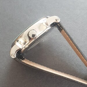 Puede incluir: Un reloj de pulsera plateado con una correa de cuero negro. El reloj tiene una esfera redonda y una correa plateada.