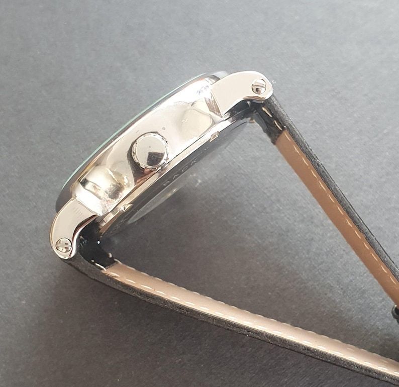 Puede incluir: Un reloj de pulsera plateado con una correa de cuero negro. El reloj tiene una esfera redonda y una correa plateada.