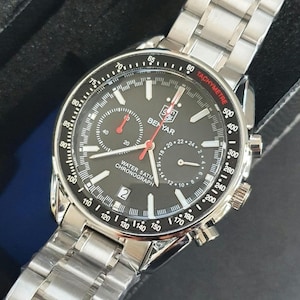 Op de afbeelding: Een zilverkleurig chronograaf polshorloge met een zwarte wijzerplaat en rode accenten. Het horloge heeft een tachymeterschaal rond de buitenrand van de wijzerplaat en de woorden "WATER 5ATM CHRONOGRAPH" gedrukt op de wijzerplaat. Het horloge heeft een zilveren metalen band.