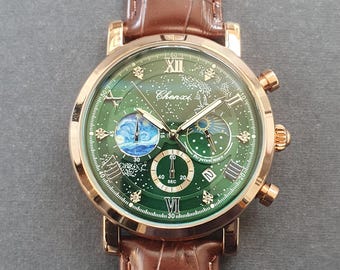 Puede incluir: Un reloj de pulsera dorado con esfera verde y correa de cuero marrón. La esfera del reloj presenta un diseño celestial detallado con la marca "Chenxi". La correa tiene una textura de piel de cocodrilo.