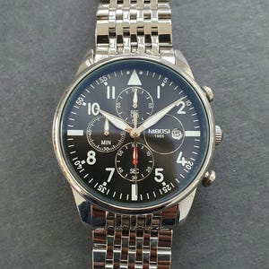 May include: A silver chronograph wristwatch with a black face and silver hands. The watch has three subdials, one for seconds, one for minutes, and one for hours. The watch face also has the text "NIBOSI 1985" and the number "15" in a red circle.