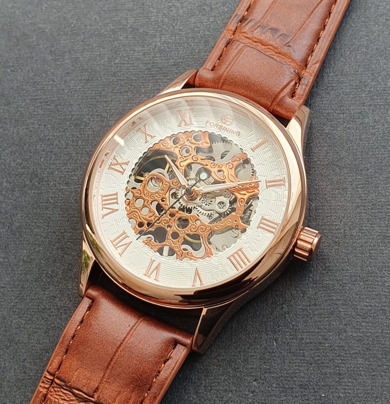 Pode incluir: Um rel&oacute;gio esqueleto de ouro rosa com marcadores de hora em algarismos romanos e uma pulseira de couro marrom. O mostrador do rel&oacute;gio &eacute; branco com um movimento mec&acirc;nico vis&iacute;vel.