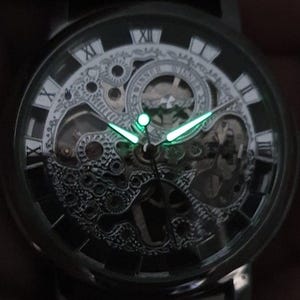 May include: A black and silver skeleton watch with Roman numeral hour markers and luminous green hands. The watch face is transparent, revealing the intricate inner workings of the watch.