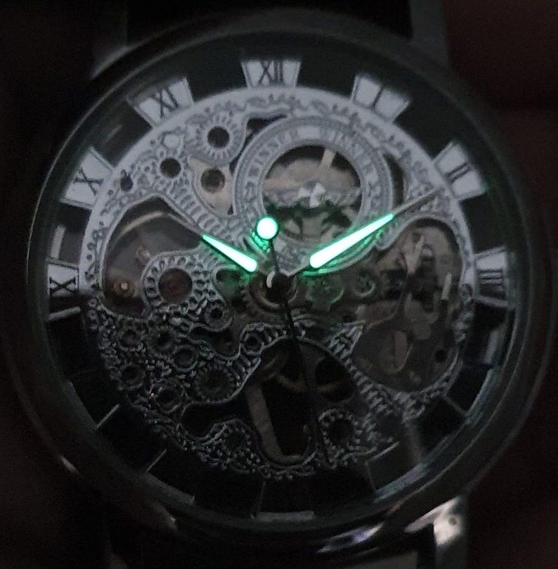 May include: A black and silver skeleton watch with Roman numeral hour markers and luminous green hands. The watch face is transparent, revealing the intricate inner workings of the watch.