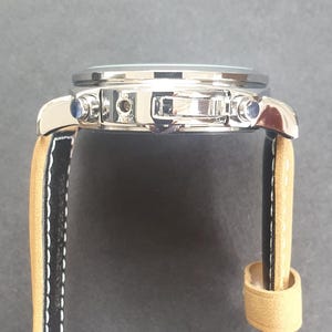 May include: A silver-toned wristwatch with a black leather band and a tan leather strap. The watch has a chronograph function and a date window.