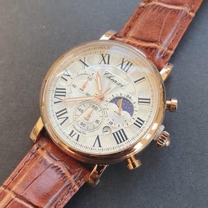 May include: A gold-tone analog wristwatch with a white face and Roman numeral hour markers. The watch has a brown leather band and features a chronograph, moon phase, and date display. The watch face also includes the text "Chenxi" and "Chronometer".