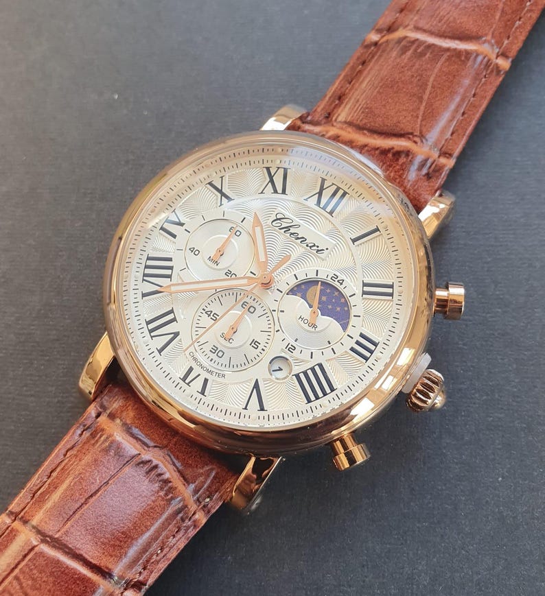 May include: A gold-tone analog wristwatch with a white face and Roman numeral hour markers. The watch has a brown leather band and features a chronograph, moon phase, and date display. The watch face also includes the text "Chenxi" and "Chronometer".