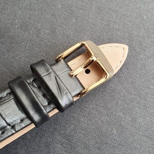 May include: Close-up of a black leather watch strap with a gold buckle. The strap has a crocodile-embossed pattern.