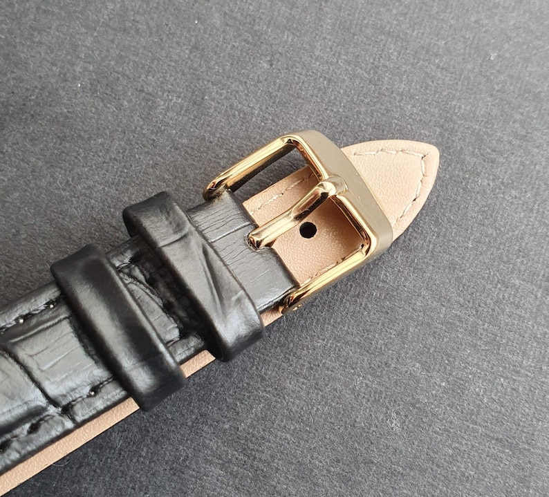 May include: Close-up of a black leather watch strap with a gold buckle. The strap has a crocodile-embossed pattern.