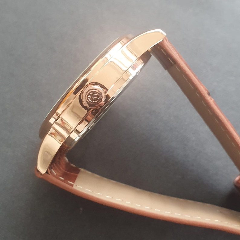 Pode incluir: Um rel&oacute;gio de pulso de ouro rosa com uma pulseira de couro marrom. O mostrador do rel&oacute;gio &eacute; vis&iacute;vel e tem um pequeno logotipo na lateral.