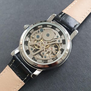 May include: A silver-toned wristwatch with a black leather band. The watch face is clear, revealing the intricate inner workings of the mechanical movement.