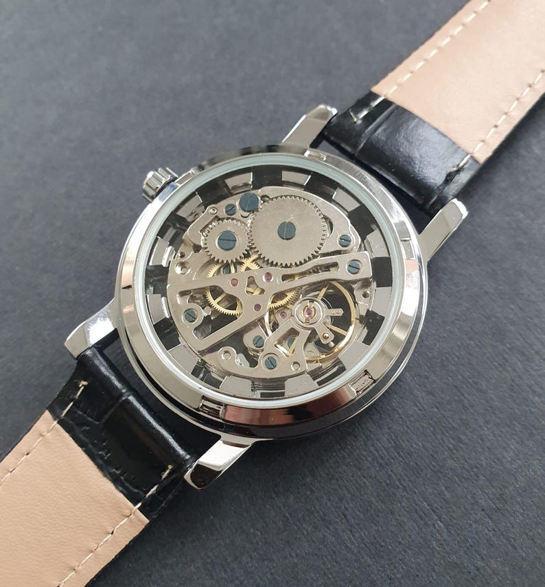 May include: A silver-toned wristwatch with a black leather band. The watch face is clear, revealing the intricate inner workings of the mechanical movement.