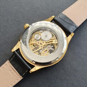May include: A gold-tone watch with a black leather band. The watch face is clear, revealing the inner workings of the mechanical movement. The watch face is labeled "CAL AUTOMATIC MECH."
