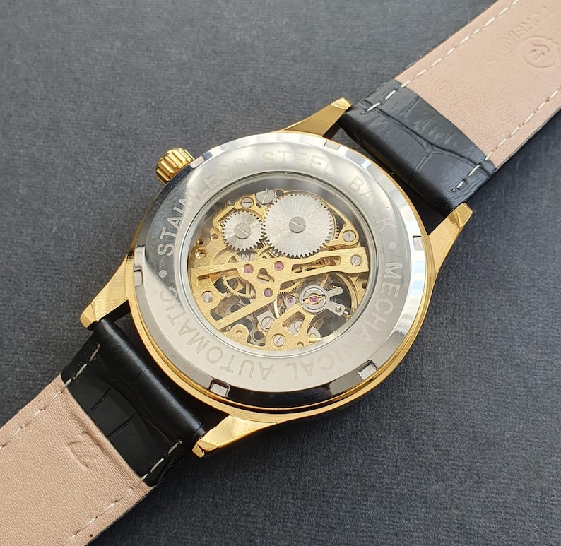 May include: A gold-tone watch with a black leather band. The watch face is clear, revealing the inner workings of the mechanical movement. The watch face is labeled "CAL AUTOMATIC MECH."