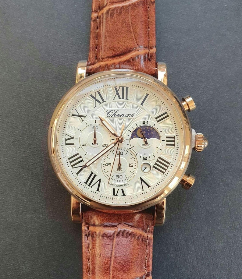 May include: A gold-tone analog wristwatch with a brown leather band. The watch face has Roman numeral hour markers and a chronograph with three subdials. The watch face also features a moon phase indicator. The brand name "Chenxi" is printed on the watch face.