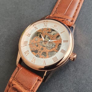 Pode incluir: Um rel&oacute;gio esqueleto de ouro rosa com uma pulseira de couro marrom. O mostrador do rel&oacute;gio tem marcadores de hora com algarismos romanos e um movimento mec&acirc;nico vis&iacute;vel.