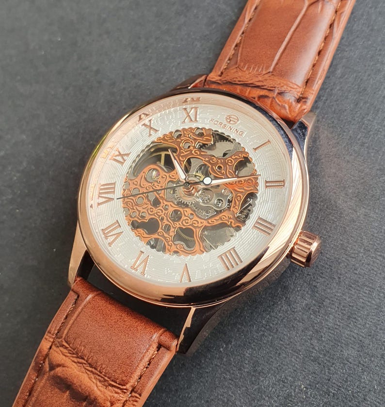 Pode incluir: Um rel&oacute;gio esqueleto de ouro rosa com uma pulseira de couro marrom. O mostrador do rel&oacute;gio tem marcadores de hora com algarismos romanos e um movimento mec&acirc;nico vis&iacute;vel.