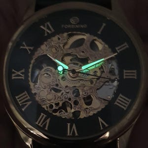May include: A gold-tone watch with a black face and Roman numeral hour markers. The watch has a skeletonized design, revealing the inner workings of the movement. The hands and hour markers glow in the dark.