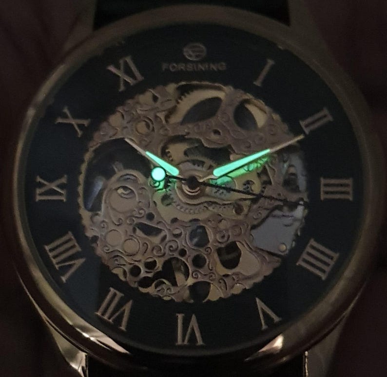 May include: A gold-tone watch with a black face and Roman numeral hour markers. The watch has a skeletonized design, revealing the inner workings of the movement. The hands and hour markers glow in the dark.