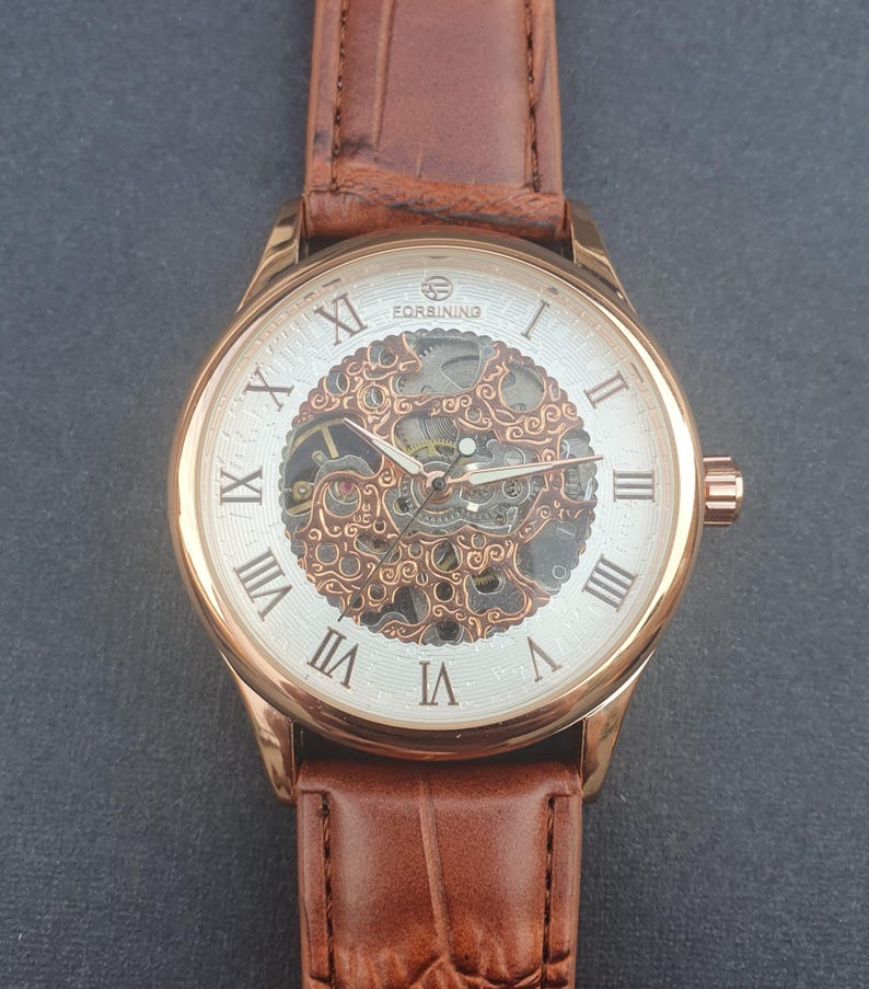 Pode incluir: Um rel&oacute;gio dourado com uma pulseira de couro marrom. O mostrador do rel&oacute;gio &eacute; branco com marcadores de hora em algarismos romanos e um movimento esquel&eacute;tico. O mostrador do rel&oacute;gio tamb&eacute;m tem o texto "FORSINING" nele.