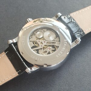 Puede incluir: Un reloj plateado con una correa de cuero negro. El reloj tiene una parte posterior transparente que revela el funcionamiento interno del reloj. La esfera del reloj tiene el texto "CASE STEEL" y "STAINLESS SS" impreso en &eacute;l.