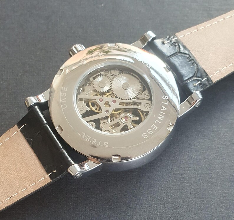 Puede incluir: Un reloj plateado con una correa de cuero negro. El reloj tiene una parte posterior transparente que revela el funcionamiento interno del reloj. La esfera del reloj tiene el texto "CASE STEEL" y "STAINLESS SS" impreso en &eacute;l.