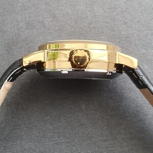 May include: A gold-tone wristwatch with a black leather band. The watch has a round face and a gold-tone bezel.