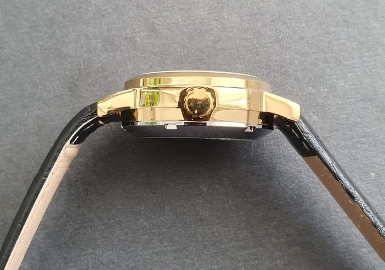 May include: A gold-tone wristwatch with a black leather band. The watch has a round face and a gold-tone bezel.