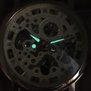 May include: A close-up of a silver skeleton watch face with luminous green hands and hour markers. The watch face is surrounded by a black bezel and a black leather strap.