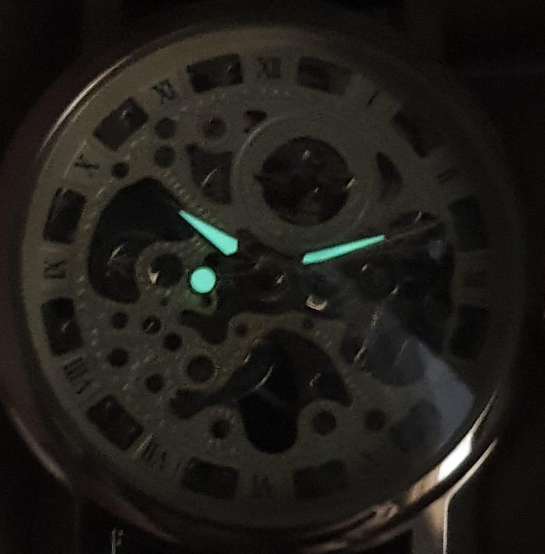 May include: A close-up of a silver skeleton watch face with luminous green hands and hour markers. The watch face is surrounded by a black bezel and a black leather strap.