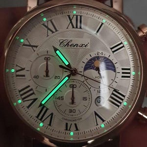 May include: A white analog watch with Roman numeral hour markers and a black and white moon phase dial. The watch face has a green glow-in-the-dark feature. The watch face also has the text "Chenxi" and "Chronometer".