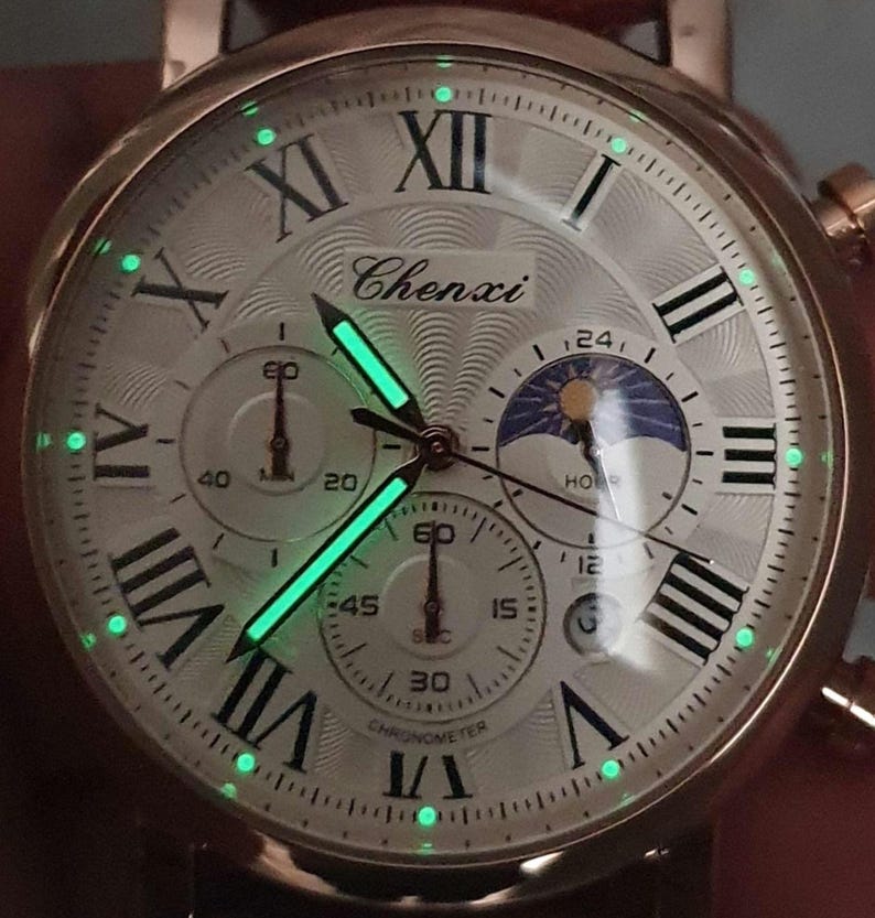 May include: A white analog watch with Roman numeral hour markers and a black and white moon phase dial. The watch face has a green glow-in-the-dark feature. The watch face also has the text "Chenxi" and "Chronometer".