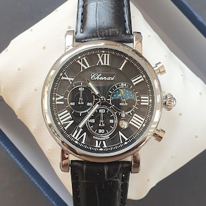 Puede incluir: Un reloj de pulsera cronógrafo plateado con una correa de cuero negro. La esfera del reloj tiene marcadores de hora en números romanos y un fondo negro. La esfera del reloj también presenta un indicador de fase lunar y la palabra "Chenxi" impresa en la esfera.