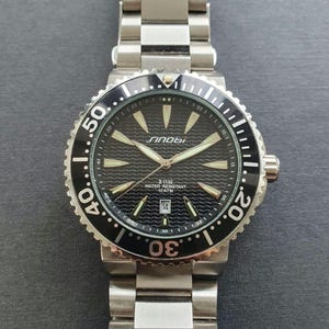 Op de afbeelding: Een zwart en zilveren herenhorloge met een zwarte wijzerplaat en een zilveren band. Het horloge heeft een datumvenster op de 3 uur positie en een zwarte bezel met witte markeringen. De wijzerplaat van het horloge heeft een golfpatroon en de tekst "Sinobu II-1132 Water Resistant 10 ATM" is op de wijzerplaat gedrukt.
