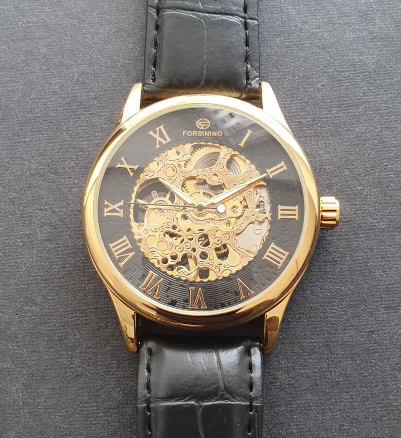 May include: A gold-tone watch with a black face and Roman numeral hour markers. The watch has a skeletonized movement, meaning the inner workings of the watch are visible. The watch is on a black leather band.