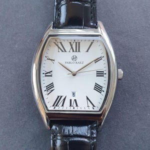 Könnte beinhalten: Eine silberne Uhr mit weißem Zifferblatt und schwarzen römischen Ziffern als Stundenmarkierungen. Die Uhr hat ein schwarzes Lederarmband und trägt die Aufschrift "PABLO RAEZ" auf dem Zifferblatt.