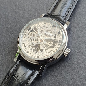 Könnte beinhalten: Eine silberne Skelettuhr mit einem schwarzen Lederarmband. Das Zifferblatt der Uhr hat römische Ziffern für die Stunden und ein transparentes Fenster, das das Innenleben der Uhr zeigt.