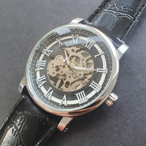 Puede incluir: Un reloj de pulsera plateado con una esfera negra y marcadores de hora en n&uacute;meros romanos. El reloj tiene una parte posterior transparente que revela el funcionamiento interno del movimiento mec&aacute;nico. El reloj est&aacute; en una correa de cuero negra.